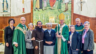Smiling clergy and lay people pose in a church before an altar, crucifix, and Marian statue, with a bishop holding a staff
