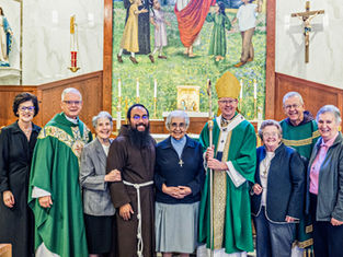 Smiling clergy and lay people pose in a church before an altar, crucifix, and Marian statue, with a bishop holding a staff