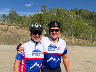 Two smiling cyclists in Samaritan Cycling jerseys and helmets pose on a roadside with forested hills under a blue sky.
