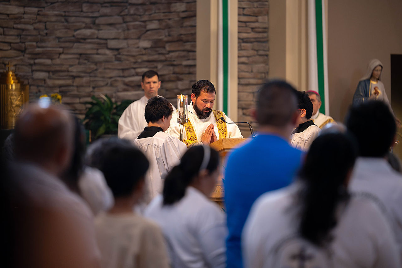 Priest in gold vestments prays at a church lectern while altar servers and congregation stand in quiet worship.