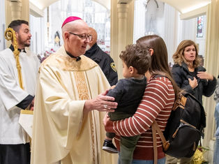 Catholic bishop in white and pink vestments blesses a mother holding a child in a church, with people gathered around them.