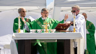 Bishop in green vestments celebrates outdoor Mass at altar under white tents, with assistants and gold chalices.