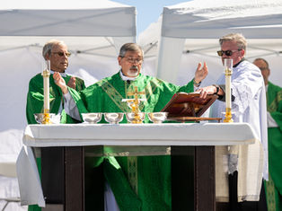 Bishop in green vestments celebrates outdoor Mass at altar under white tents, with assistants and gold chalices.
