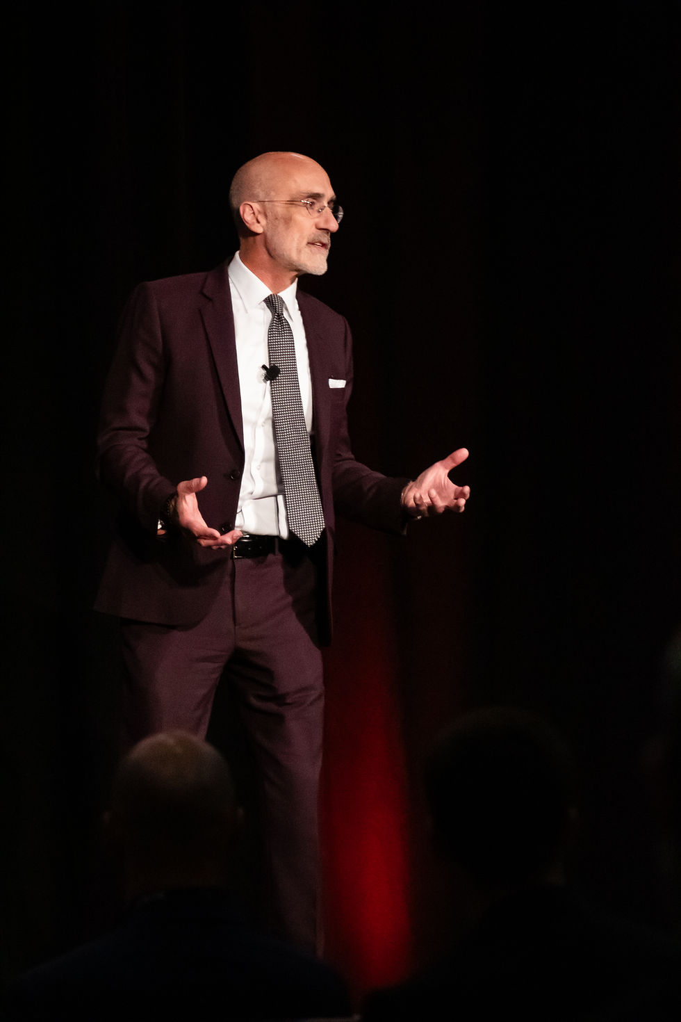 Bald man in burgundy suit speaks on a dark stage, gesturing with open hands under red lighting.