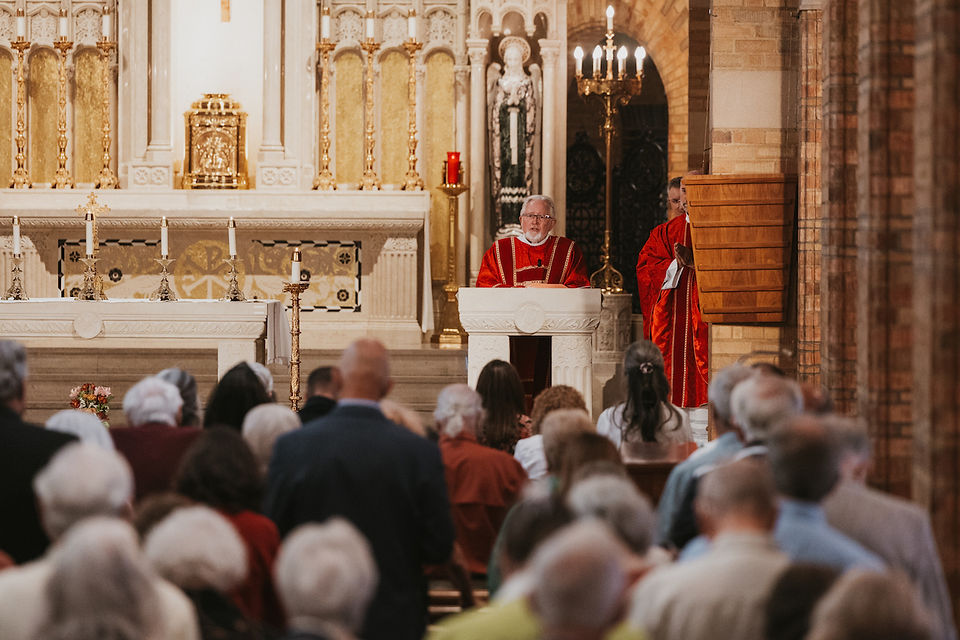 Priest in red vestments speaks at an ornate church altar while a congregation stands listening in the pews.