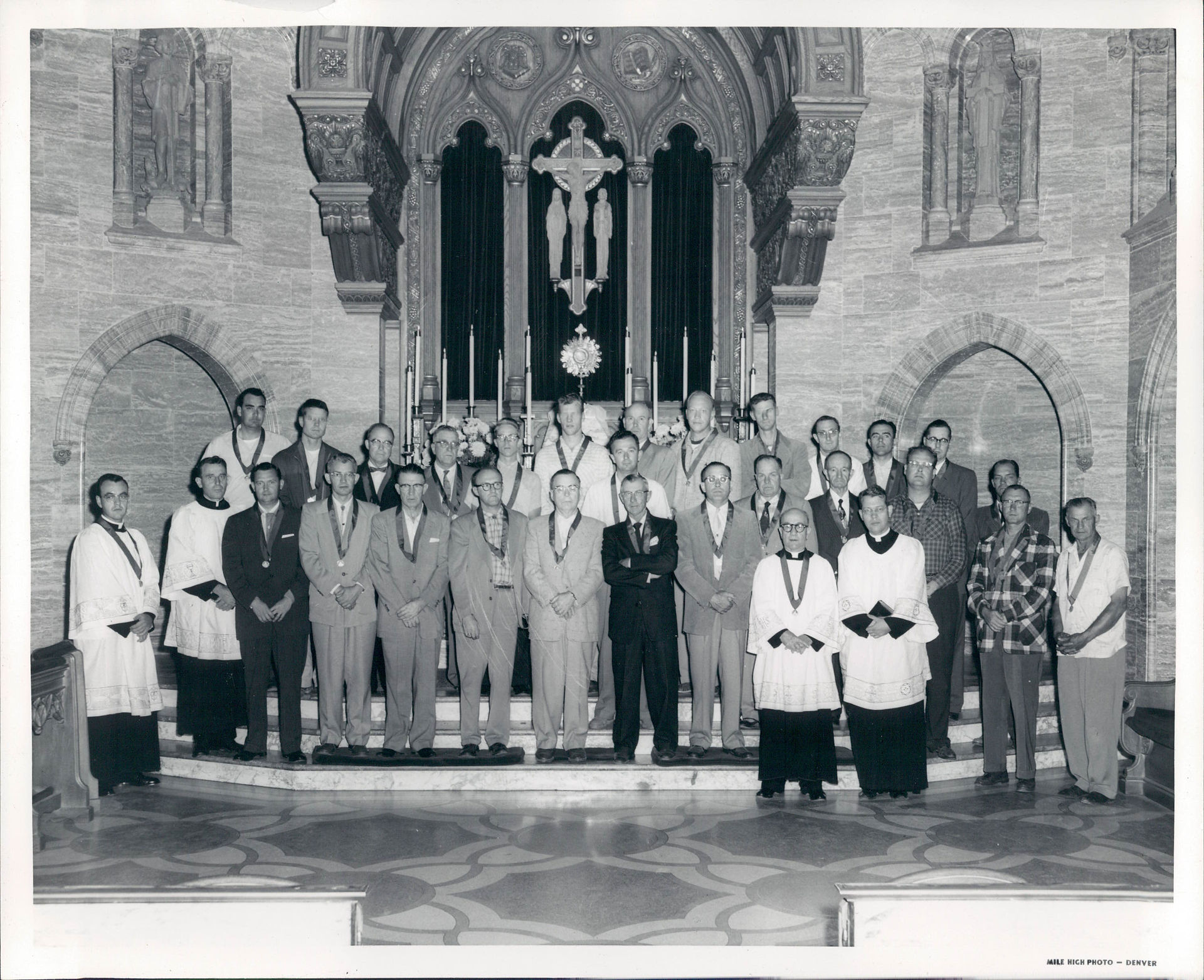 Black-and-white group photo of men posing at a church altar beneath a large cross, with ornate arches and solemn mood.