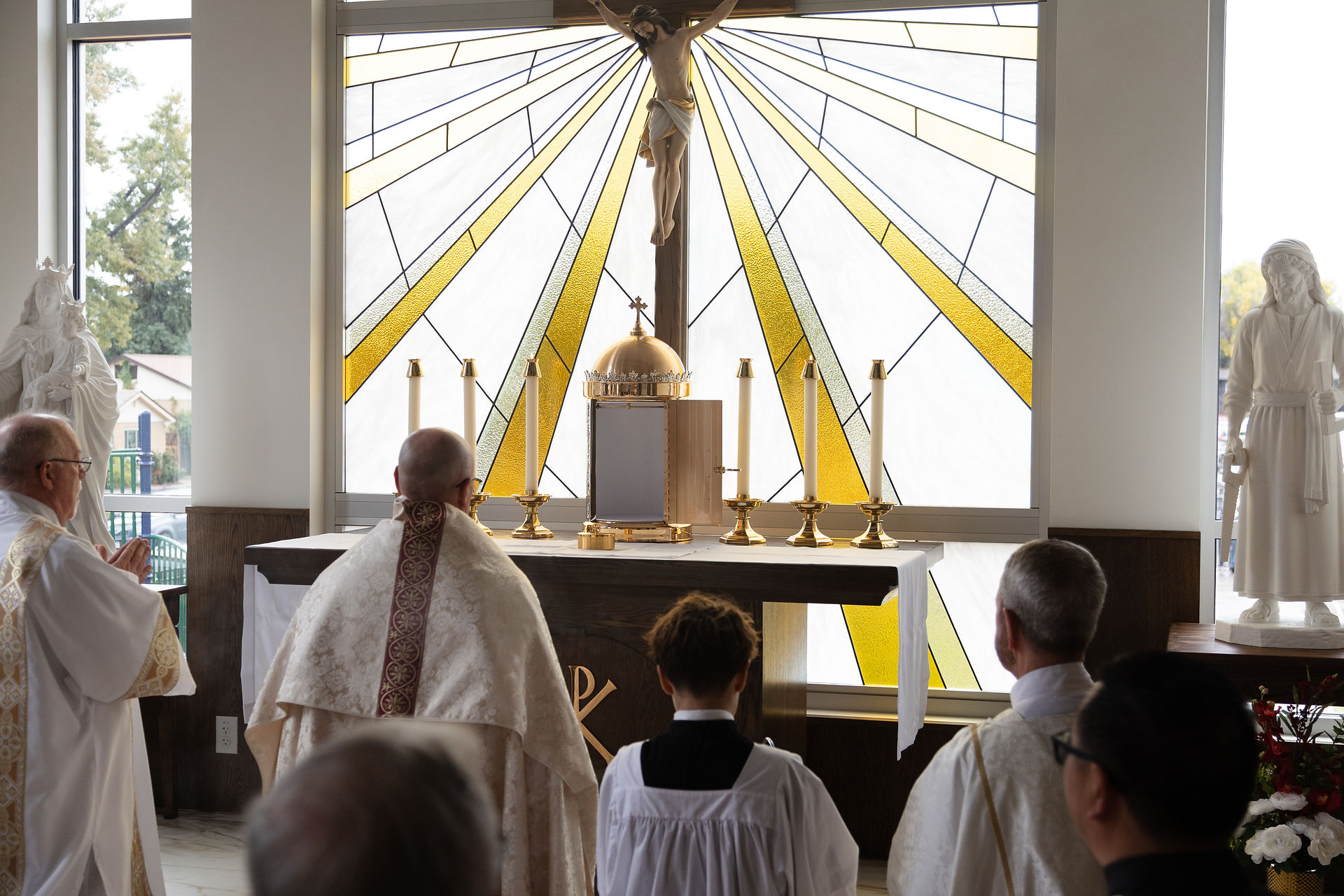 Priests and worshippers kneel at a Catholic altar with crucifix, gold tabernacle, candles, and stained glass in a quiet chapel.
