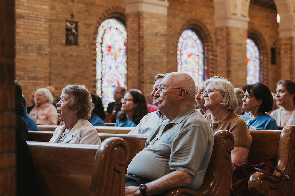 Elderly congregation seated in wooden church pews, listening attentively beneath stained-glass windows in a warm brick interior.