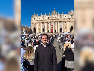 Smiling priest in black cassock stands before a seated crowd at St. Peter’s Basilica under a clear blue sky.