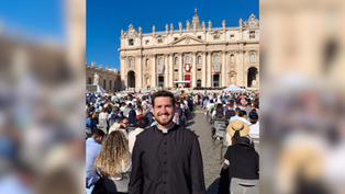 Smiling priest in black cassock stands before a seated crowd at St. Peter’s Basilica under a clear blue sky.