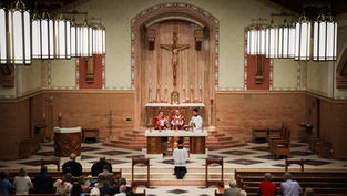 Clergy celebrate Mass at a church altar beneath a crucifix, with worshippers standing in the pews under glowing pendant lights.