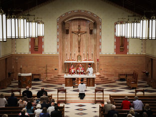 Clergy celebrate Mass at a church altar beneath a crucifix, with worshippers standing in the pews under glowing pendant lights.