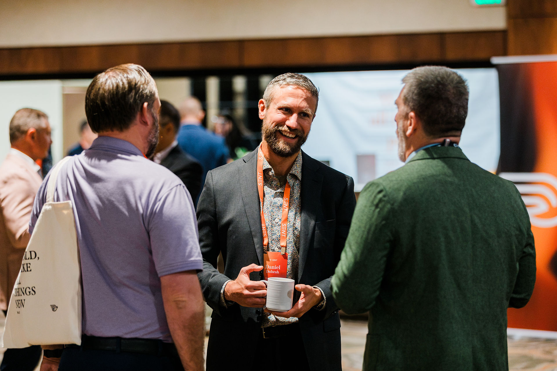 Smiling men chat at a conference; one in a suit with a Daniel Dobesh lanyard holds a mug in a busy indoor lobby.