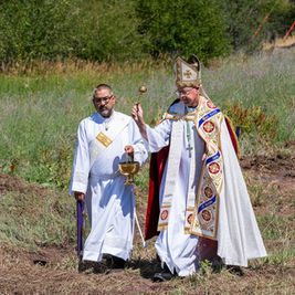 Two Catholic clergy in white vestments walk through a grassy field, one holding a chalice and the other a bishop’s staff.