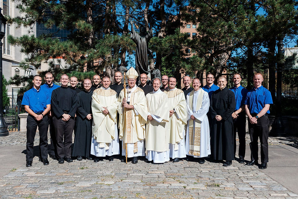 Group of priests and seminarians pose outdoors before a statue and pine trees, with one bishop in white at center.