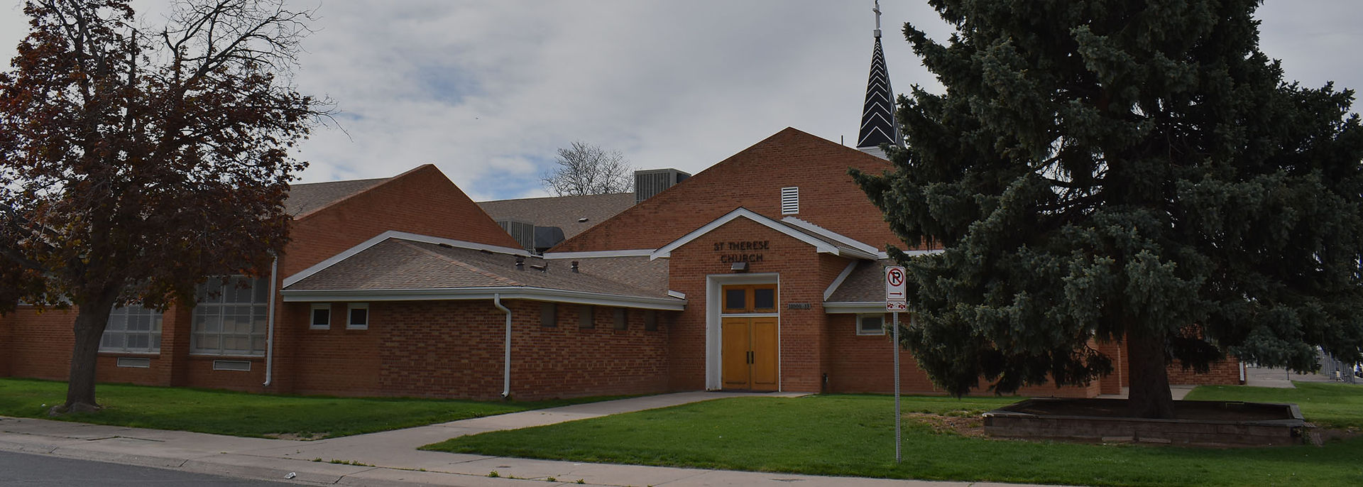 Brick St. Therese Church with steeple, framed by trees and lawn under cloudy sky; no parking sign by the entrance