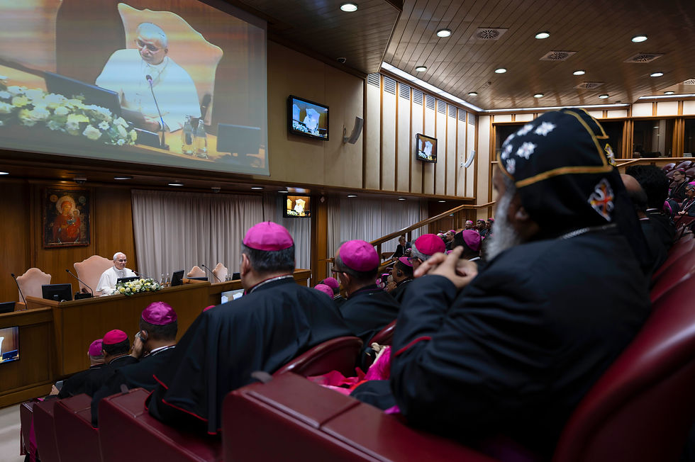 Catholic bishops in black robes and magenta caps watch a pope speaking on large screens in a wood-paneled auditorium.