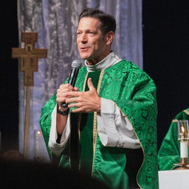 Priest in green vestments speaks into a microphone during a church service, with another bishop and altar candles behind him.