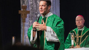 Priest in green vestments speaks into a microphone during a church service, with another bishop and altar candles behind him.