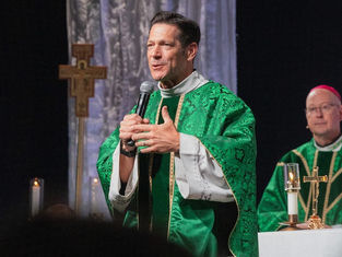 Priest in green vestments speaks into a microphone during a church service, with another bishop and altar candles behind him.