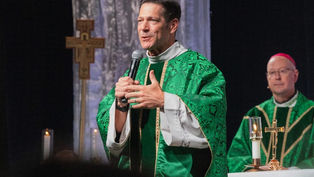 Priest in green vestments speaks into a microphone during a church service, with another bishop and altar candles behind him.