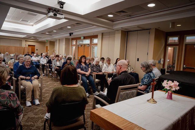 Group of older adults seated in a hotel meeting room, listening in a circle near a table with flowers and a black stage.