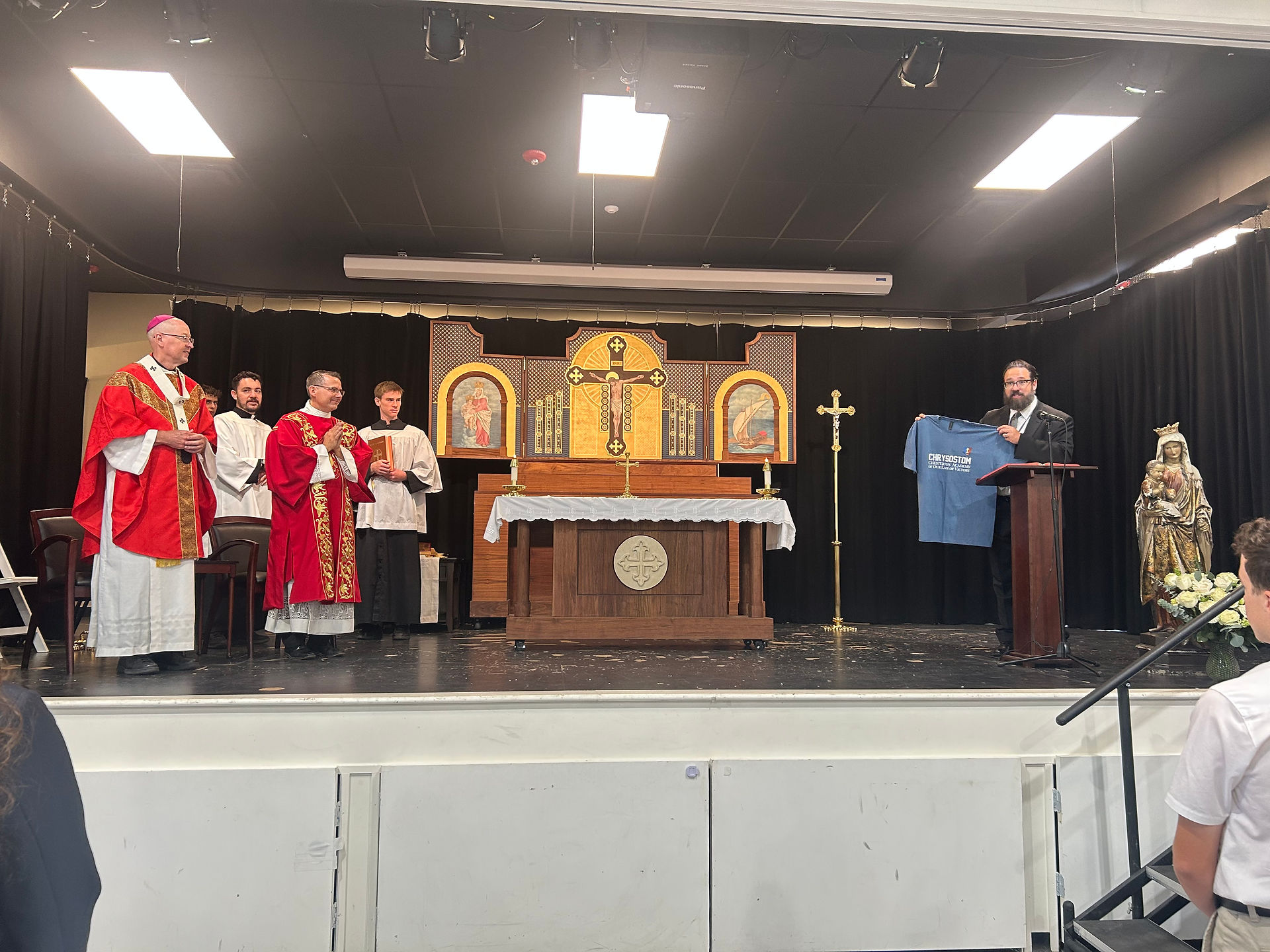 Clergy on a church stage beside an altar; a man at a podium holds up a blue Chrysostom Christian Academy shirt with text.