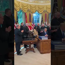 Donald Trump bows his head at the Resolute Desk in the Oval Office while bishops pray over him among U.S. flags and gold drapes.