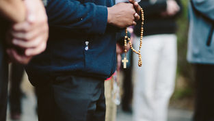 Close-up of a person holding rosary beads and a paper during an outdoor prayer gathering, with others blurred in background.