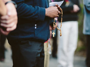 Close-up of a person holding rosary beads and a paper during an outdoor prayer gathering, with others blurred in background.