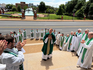 Clergy in white and green vestments clap around a smiling priest outside a university fountain and brick campus gate.