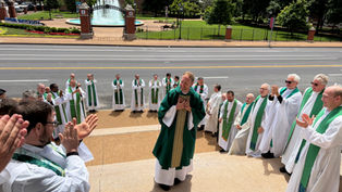Clergy in white and green vestments clap around a smiling priest outside a university fountain and brick campus gate.