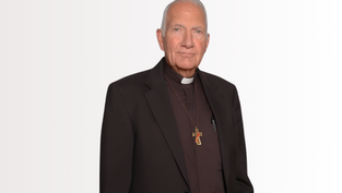 Elderly deacon in black clerical suit with cross pendant stands against a plain white background, looking calmly at the camera.