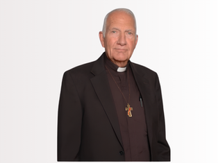 Elderly deacon in black clerical suit with cross pendant stands against a plain white background, looking calmly at the camera.