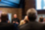 Bald priest in black clerical suit speaks at a podium to an audience in a brick-walled church hall.