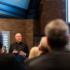Bald priest in black clerical suit speaks at a podium to an audience in a brick-walled church hall.