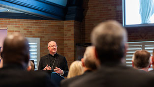 Bald priest in black clerical suit speaks at a podium to an audience in a brick-walled church hall.
