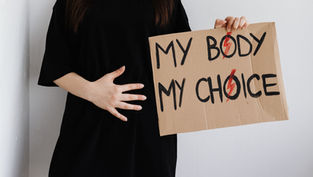 Person in black holds a cardboard sign reading MY BODY MY CHOICE with red lightning marks, against a white wall.