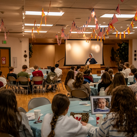Nun speaks at a podium to seated teens in a hall decorated with hanging U.S. and Spanish flags, during a warm community gathering.