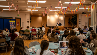 Nun speaks at a podium to seated teens in a hall decorated with hanging U.S. and Spanish flags, during a warm community gathering.