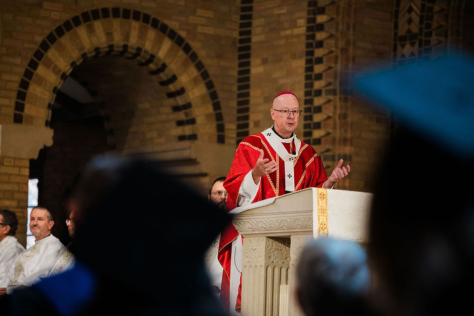 Bishop in red vestments speaks at a church pulpit, gesturing to a blurred congregation beneath arched brick walls.