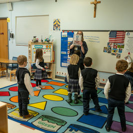 Teacher holds up a picture book to a circle of young students in a colorful classroom with a whiteboard, flag, and crucifix.