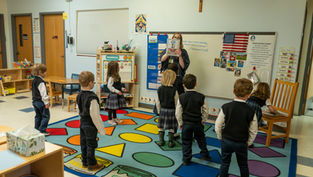 Teacher holds up a picture book to a circle of young students in a colorful classroom with a whiteboard, flag, and crucifix.