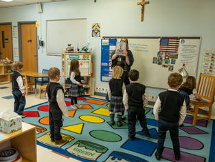Teacher holds up a picture book to a circle of young students in a colorful classroom with a whiteboard, flag, and crucifix.