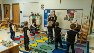 Teacher holds up a picture book to a circle of young students in a colorful classroom with a whiteboard, flag, and crucifix.