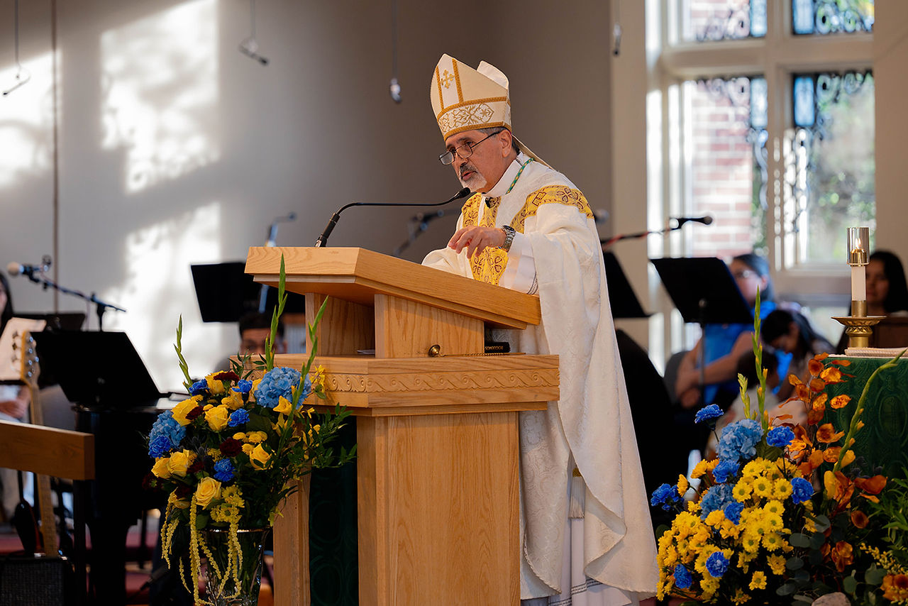 Bishop in white and gold vestments speaks at a wooden pulpit in a sunlit church, with flowers and musicians behind him.