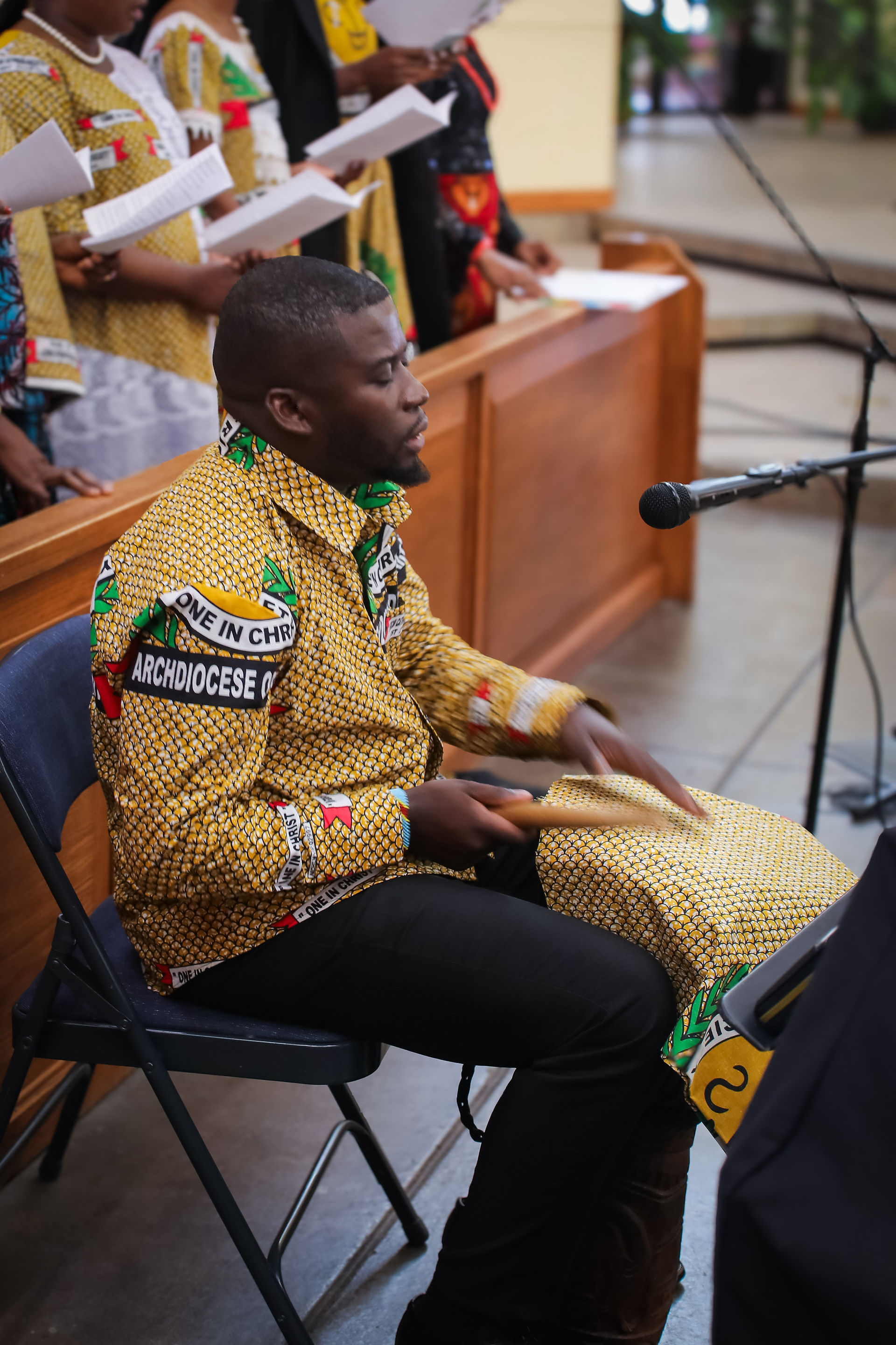 Man in patterned shirt drums and sings at church with choir behind him; shirt reads ONE IN CHRIST and ARCHDIOCESE.