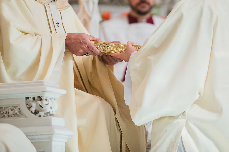 Bishop in white vestments passes a gilded ceremonial object during a solemn church service.