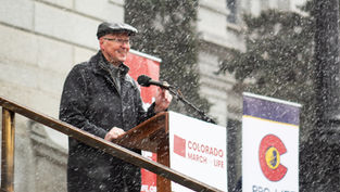 Man in black coat and cap speaks at a podium in falling snow, beside Colorado March for Life and Pro-Life Colorado signs.