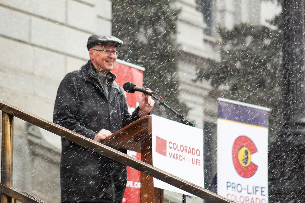 Man in black coat and cap speaks at a podium in falling snow, beside Colorado March for Life and Pro-Life Colorado signs.